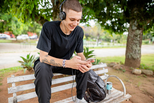Smiling young man wearing headphones streaming a video on his smart phone while sitting on a park bench