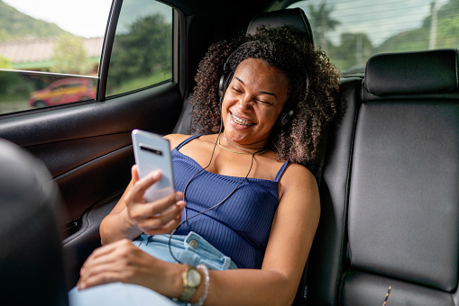 Young woman wearing headphones laughing at a video ono her smart phone while riding in the back of a taxi