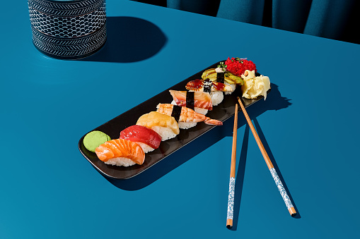 Sushi set featuring salmon, tuna, shrimp, eel, and avocado on a black plate against a deep blue backdrop with chopsticks. Dramatic lighting and shadows present. Perfect for an Asian restaurant menu