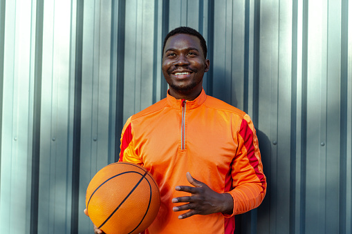 Smiling young African American man standing in front of the gray background and holding basketball ball