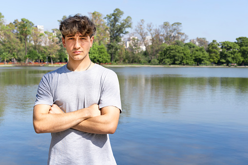 Portrait of confident young blond man with crossed arms looking at camera, summer lake background. Copy space