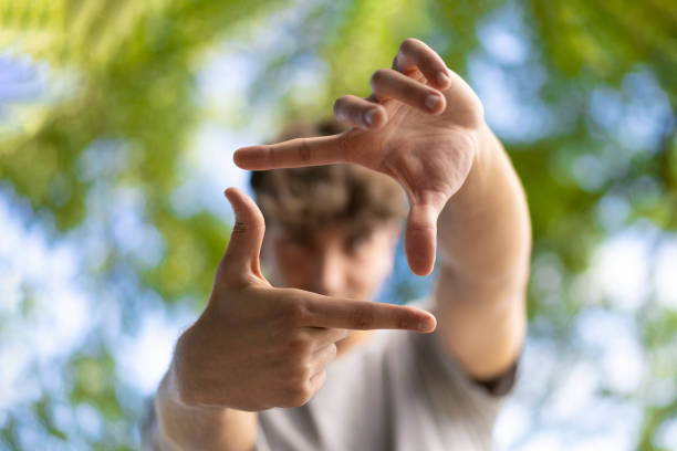 A handsome young man blinks his eyes and looks through a frame made up of hands. Attractive man make frame by fingers, isolated on blur background with copy space for text.