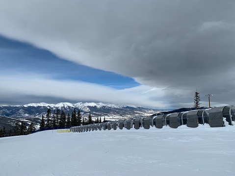This striking photograph captures a tranquil winter scene in the heart of the Rockies. Nestled in the foreground is a snow-covered ski lift belt, a silent sentinel in the midst of a serene snowscape. Above, the majesty of the mountain range comes into view, their snowy peaks emerging from a uniquely curved cloud formation. It’s a captivating blend of man-made and natural elements, a testament to the power and beauty of the great outdoors.