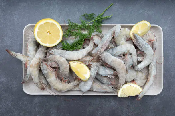 Fresh shrimp is scattered on the table with lemon slices. Products for grilling. Lemon slices and frozen shrimp.
