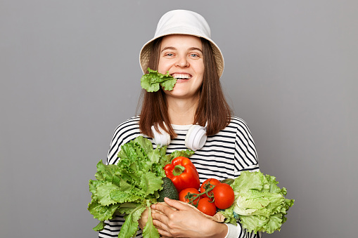 Happy positive woman holding vegetables isolated over gray background biting lettuce keeps green diet feel healthy enjoying organic nutrition.