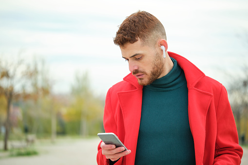 Portrait of latin young man with a red coat and wireless headphones listening music in the street in autumn.