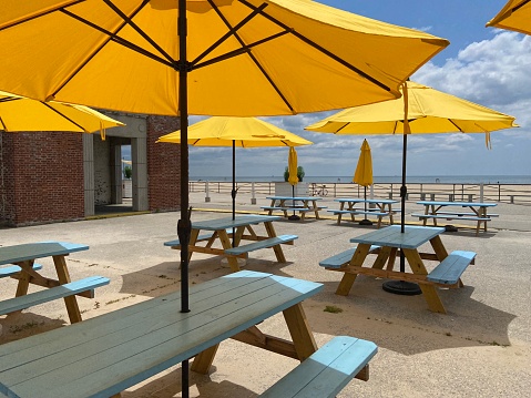 Painted blue picnic tables and yellow sun umbrellas arranged in a row in front of the concession stands at Riis Beach Cooperative at Jacob Riis Park in the Rockaways in Queens, New York