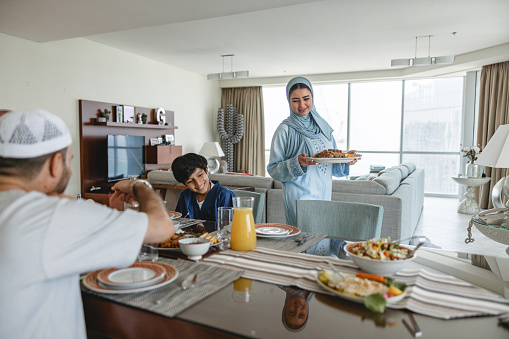 Middle eastern mother bringing main traditional dish to the dining table. Father and son are already sitting. Beautiful interior design in the background. They are all wearing traditional clothes.