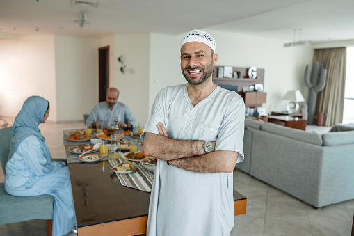 Portrait of middle eastern middle-age male smiling while standing in front of dining table. He is wearing traditional grey clothes, white muslim cap and a watch. His wife and grandfather in the background. Focus is on foreground.