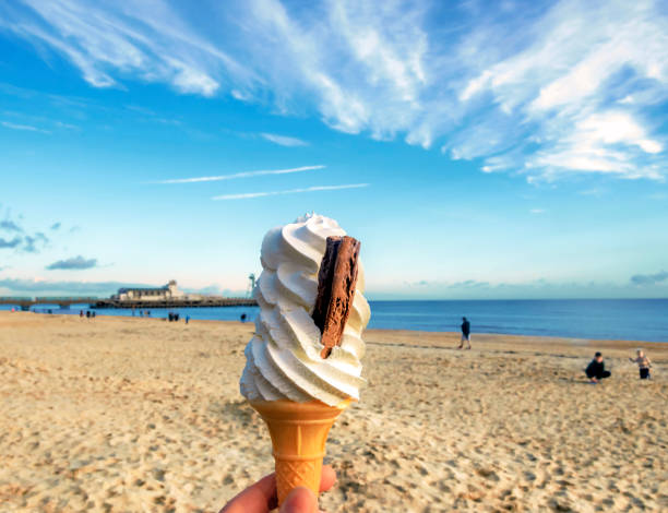 A close-up of a classic British soft whip ice cream in a cone, with a flakey type of chocolate bar pushed into the side of the ice cream. The ice cream is being held up with a view behind it in soft focus of a large stretch of sandy beach, blue sea, Bournemouth pier in the distance, and out of focus unrecognizable people on the sand. The sky is blue with whispy white clouds on a sunny day. Copy space