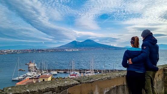 Tourist couple enjoying the panoramic view from Castel dell Ovo in city of Naples, Campania, Italy, Europe. Bathing in the sun. Panoramic view on volcano mount Vesuvius. Ferries in harbor of Naples