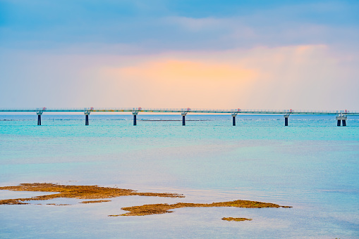 The sea with shades of blue and green in Okinawa. In the distance, a long bridge spans across the sea. The sky features scattered clouds, with a gentle ray of sunlight streaming through a gap, casting a radiant glow on the sea's surface.