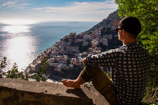 Tourist man sitting on wall enjoying panoramic view on colorful houses of coastal town Positano, Amalfi Coast, Italy, Campania, Europe. Vacation at coastline Mediterranean Sea. Path of the Gods. Awe