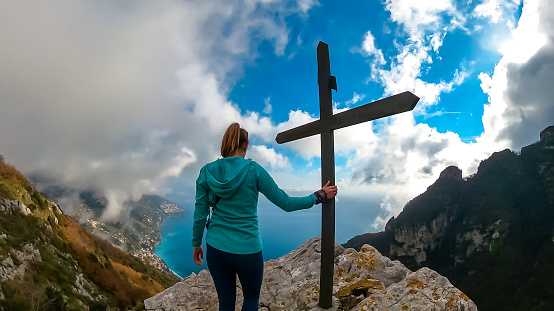 Active woman at summit cross near Santa Maria del Castello. Scenic view on coastal town Positano, Praiano. Magical hiking trail, Lattari Mountains, Apennines, Amalfi Coast, Campania, Italy, Europe