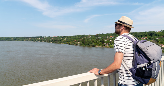 Backpacker wearing straw hat standing on the river bridge and enjoying  view of the river and nature.