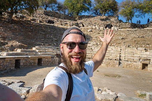Caucasian bearded man make selfie video phone call, story reel in ancient ruins greek turkish amphitheater using smartphone camera make photos while traveling in sightseeing landmark public park summer.