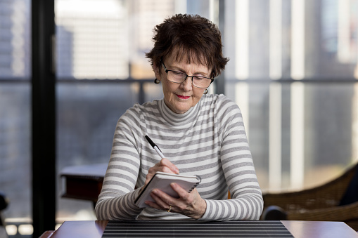 Senior woman sitting at home writing in a note pad with a pen.