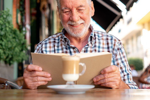 Attractive senior man sitting outdoor at a cafe table reading a book while enjoying a coffee and milk drink - caucasian elderly man relaxed in retirement or vacation