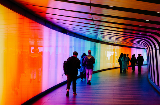 London, UK - 5 June, 2023: a crowd of people walking through an illuminated, multi-colored, futuristic underground tunnel at Kings Cross underground station in central London, UK. The tunnel is illuminated with the colours of the rainbow to mark Gay Pride month.