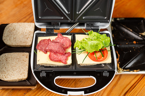 A sandwich maker with two different sandwiches with tomatoes, ham, onions and cheese. Preparing a light meal.