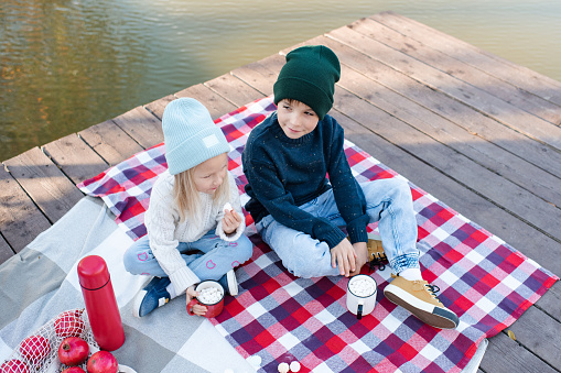 Two kids boy and girl having picnic on wooden deck over lake having fun together outdoor. Autumn season. Childhood.