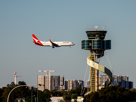 A Qantas B737-838 plane, registration VH-VXU, coming into land on the third runway of Sydney Kingsford-Smith Airport as flight QF579 from Hamilton Island.  In the foreground is the air traffic control tower.  This image was taken from Kyeemagh Avenue, Kyeemagh on a sunny winter afternoon on 10 June 2023.