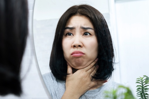 worried Asian woman checking her fat double chin under her lower jaw in front of a mirror, a sign of weight gain or obesity