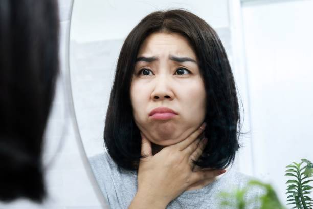 worried Asian woman checking her double chin under her lower jaw in front of a mirror, stock photo