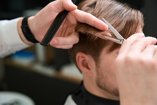 Close up on hands of barber cutting hair of his client by using scissors and comb in modern salon