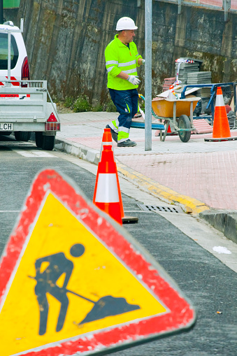 Lugo, Spain- March 30, 2022:  Road work ahead sign, urban road, danger ahead warning and road construction worker wearing reflective clothes, wheelbarrow, traffic cones. Lugo city, Galicia, Spain.