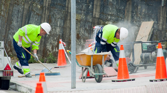 Lugo, Spain- March 30, 2022: Two road construction workers wearing reflective clothes, working,  wheelbarrow, traffic cones. Lugo city, Galicia, Spain.