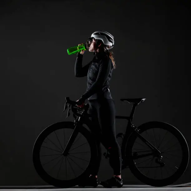 Girl posing on roadbike. Drinking from green cycling water bottle. White protective helmet. Side lit cyclist against dark background.