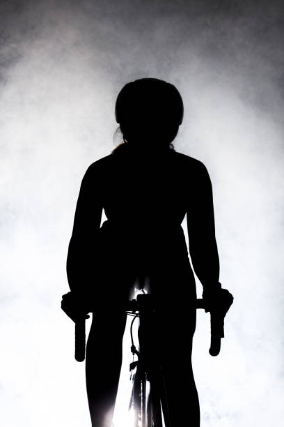 female roadbiker silhouette riding a bicycle against dark background with fog