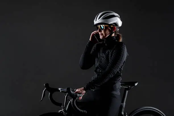Girl posing on roadbike. White protective helmet and fashion sunglasses. Talking on cell phone. Side lit cyclist against dark background.