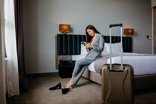 Asian Chinese businesswoman resting and using smartphone after checked in hotel room. Business travel concept.