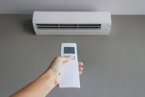 A remote control with a display showing a temperature of 20 degrees Celsius in a woman's hand in the foreground against the background of an air conditioner on a gray wall