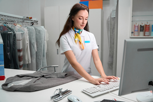 Concentrated woman laundry office operator typing on keyboard, entering data about clients garments to programm