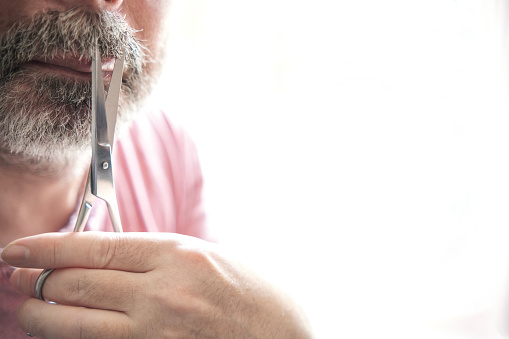 Unrecognizable adult man using scissors to cut his mustache. He has grey hair and he is wearing pink t-shirt. Back-light illuminated.