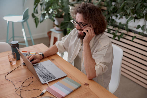 istock Smiling businessman talking by phone with client while working on laptop in cozy coworking 1497433227