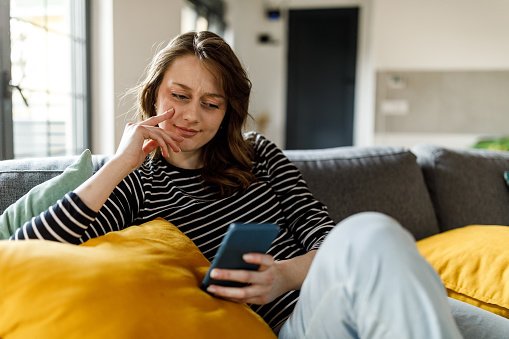 Portrait of happy young woman simply relaxing on the cozy sofa in her living room and scrolling online via smart phone.
