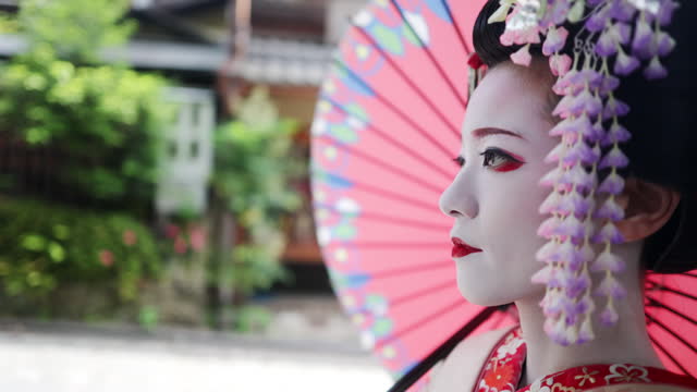 Side view of Japanese Maiko (Geisha in training) walking on street and live streaming in Gion, Kyoto - using Japanese traditional paper umbrella