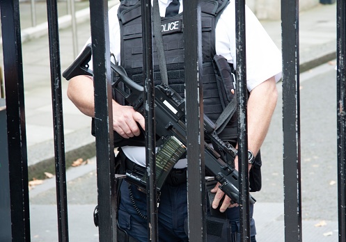 A law enforcement officer standing in a defensive position, equipped with a sidearm, in front of a chain-link fence