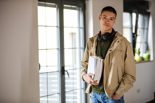 Copy space shot of charming teenage boy, headphones around his neck, standing in a room, holding school books, smiling at camera with confidence.