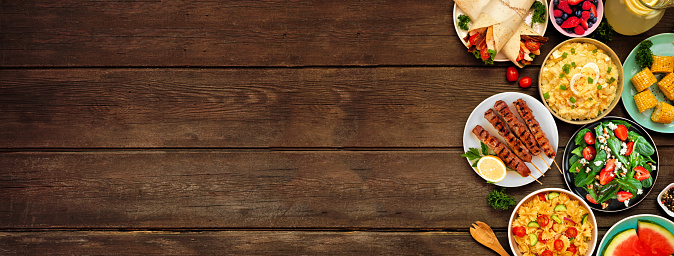 Summer food side border over a dark wood banner background. Assorted refreshing salads, fruit, wraps and BBQ grilled skewers. Overhead view. Copy space.