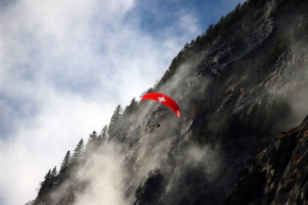 Swiss paraglider in front of dramatic steep rocky cliffs of Alps in Switzerland during lifting of fog inside Lauterbrunnen valley. Single evergreen trees on mountain ridge, blue skies and white clouds. parapente suizo frente a espectaculares acantilados rocosos de los alpes en suiza durante el levantamiento de la niebla dentro del valle de lauterbrunnen. Árboles de hoja perenne individuales en la cresta de la montaña, cielos azules y nubes blancas.