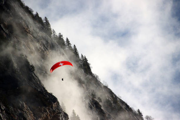 Swiss paraglider in front of dramatic steep rocky cliffs of Alps in Switzerland during lifting of fog inside Lauterbrunnen valley. Single evergreen trees on mountain ridge, blue skies and white clouds. parapente suizo frente a espectaculares acantilados rocosos de los alpes en suiza durante el levantamiento de la niebla dentro del valle de lauterbrunnen. Árboles de hoja perenne individuales en la cresta de la montaña, cielos azules y nubes blancas.