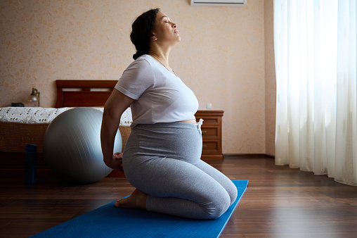 Serene conscious pregnant woman practices prenatal stretching and breathing exercises for wellness and healthy pregnancy. Side portrait of a gravid ethnic mother sitting in hero pose on a yoga mat
