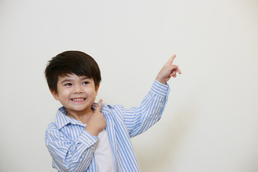 A little boy wearing a polite shirt is pointing with his hand against a white background.