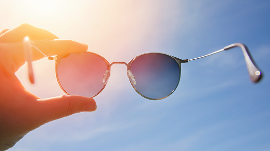 Hand holding stylish round sunglasses against bright sky and sun background. Wearing sunglasses on a sunny summer day. A man looks at the bright sun through polarized sunglasses. Summer mood