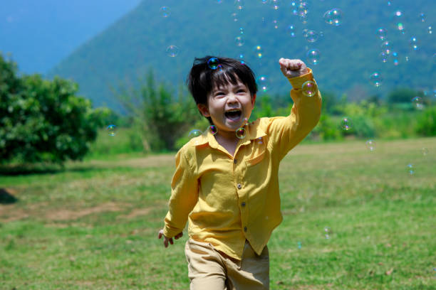 uroczy chłopiec radośnie goniący bańki mydlane w powietrzu na podwórku. - child asian and indian ethnicities facial expression asia zdjęcia i obrazy z banku zdjęć